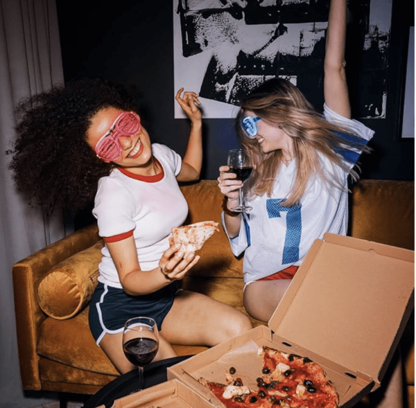 Two women sitting on a couch with pizza and wine.